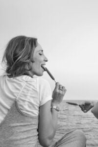 Nainen syö mehujäätä. Taustalla näkyy meri ja toisen käden pitämä mehujää. Woman is eating a popsicle with sea and someone else's hand holding another popsicle.