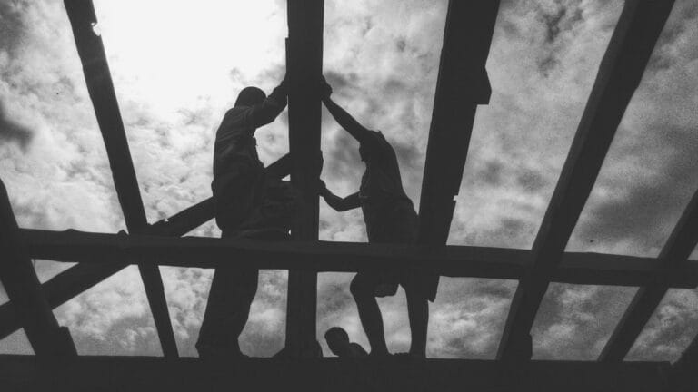 KTwo people are working on scaffolding or a roof structure, seen from above. Only their silhouettes are visible against a cloudy sky, while a third person can be seen lower down between the structures. The black-and-white photo emphasizes the contrast between light and shadow.