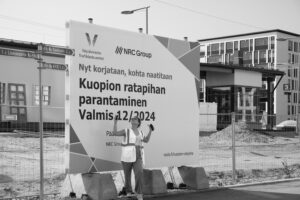 Mustavalkoinen kuva työmaasta Kuopiossa. Etualalla suuri kyltti, jossa lukee “Kuopion ratapihan parantaminen – valmis 12/2024” sekä Väyläviraston ja NRC Groupin logot. Kyltin edessä seisoo huomioliiviin pukeutunut henkilö, joka katsoo ylöspäin ja pitää kädessään puhelinta. Taustalla näkyy rakennuksia, aita sekä keskeneräinen maa-alue.